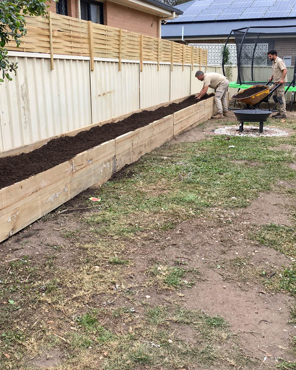 Two Ground Control landscapers filling a long timber raised garden bed with soil